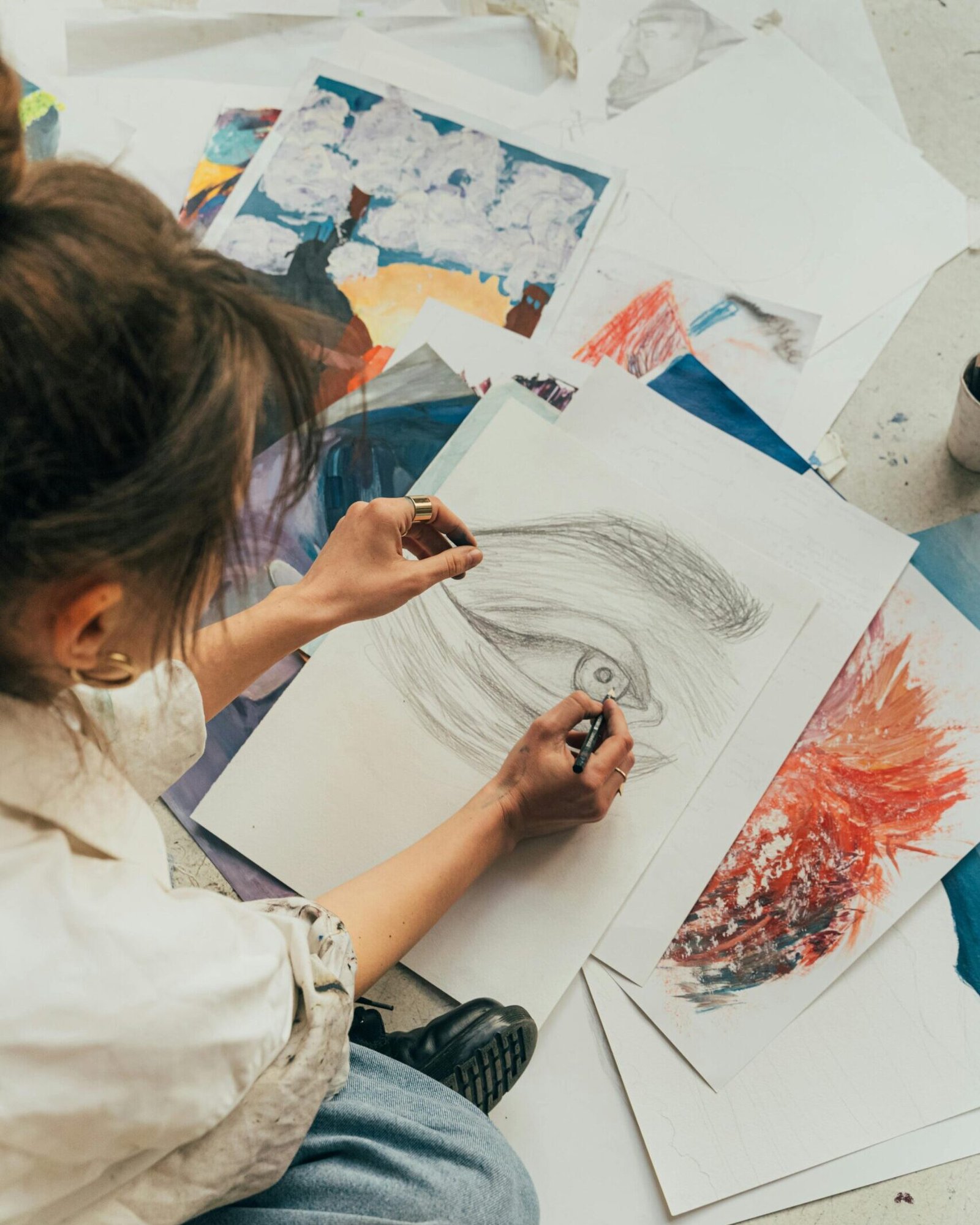 A woman sketches on the floor, surrounded by vibrant art pieces, showcasing creativity in action.